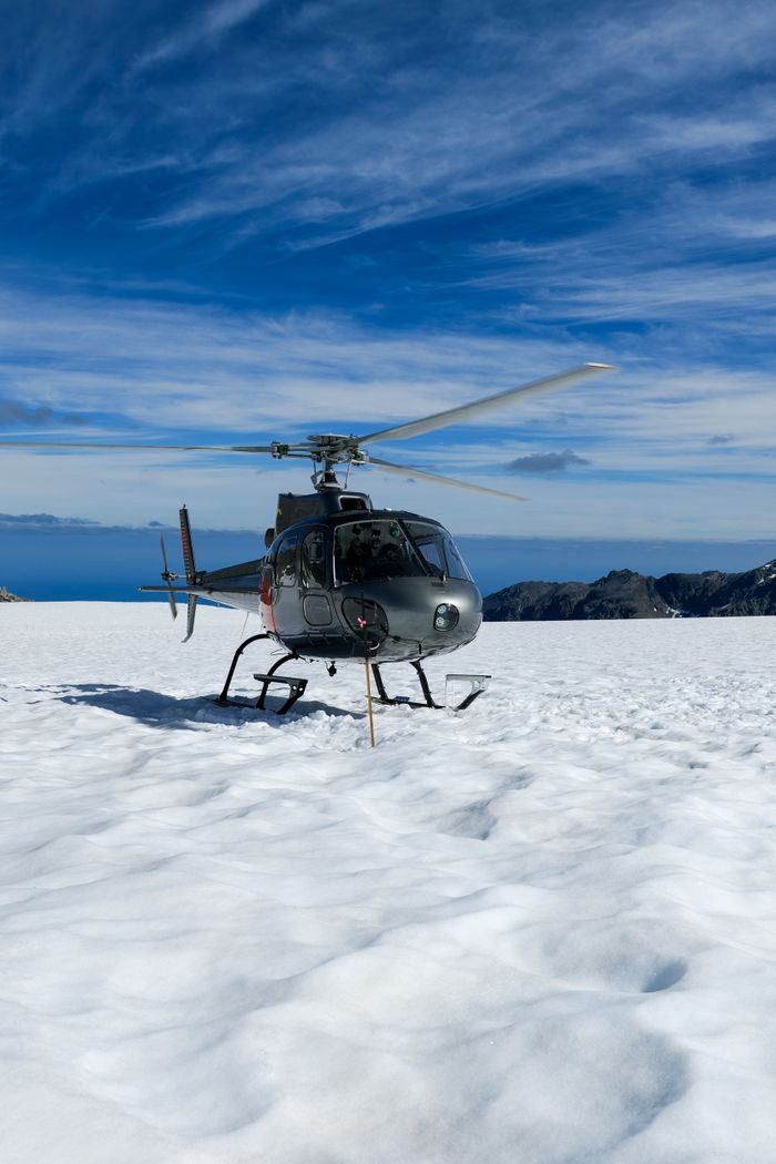 A helicopter is parked on a snow-covered mountain under a clear blue sky in New Zealand.